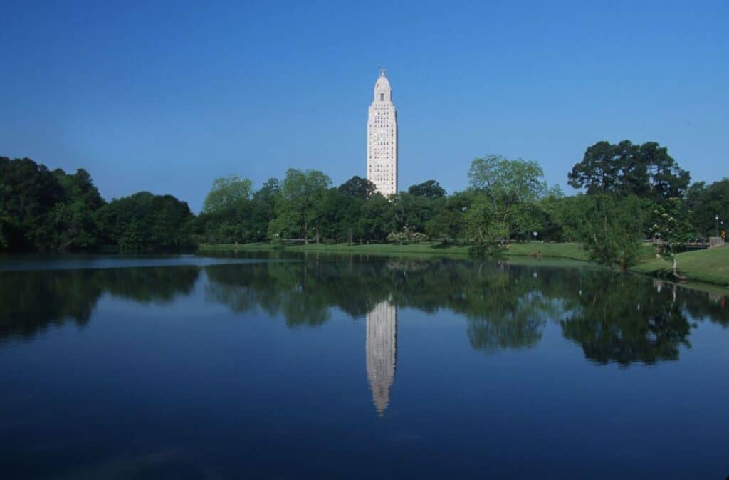 Tall white tower reflected in a calm lake, surrounded by green trees under a clear blue sky.
