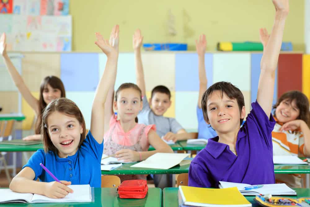 Several children in a classroom sit at desks with notebooks, raising their hands and looking toward the front of the room.