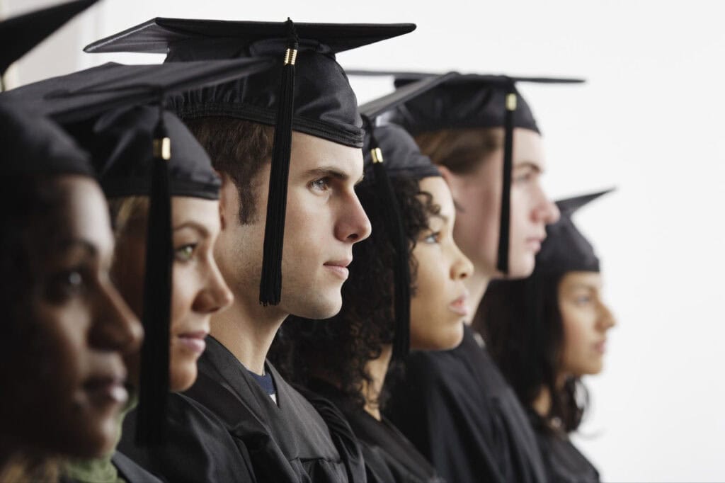 A row of young adults in graduation caps and gowns stand in profile, looking forward against a plain light background.