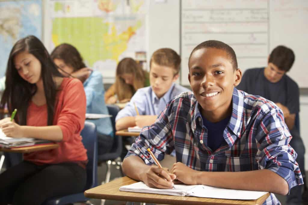 A student in a plaid shirt smiles at the camera while others write at their desks in a classroom setting.