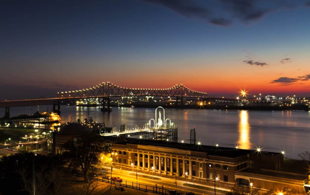 A lit bridge spans a river at dusk with city lights reflecting on the water and a building in the foreground.
