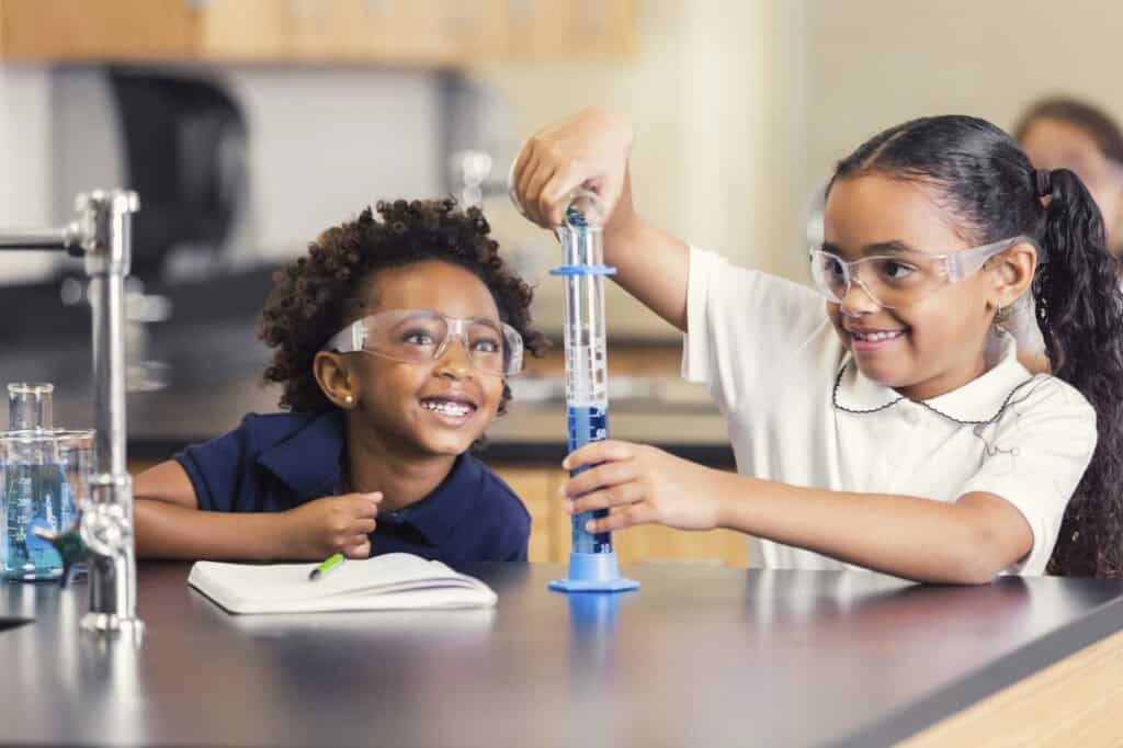 Two young girls wearing safety goggles work with a graduated cylinder of blue liquid at a science lab table, smiling.