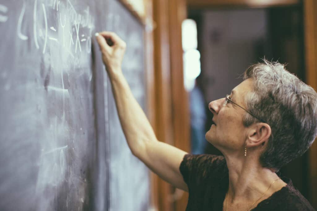 An older woman writes a math equation with chalk on a blackboard in a classroom.