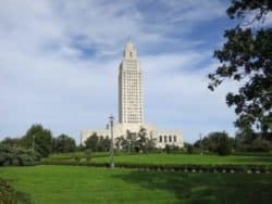 A tall, white government building stands in the distance, surrounded by a green lawn and trees under a partly cloudy sky.