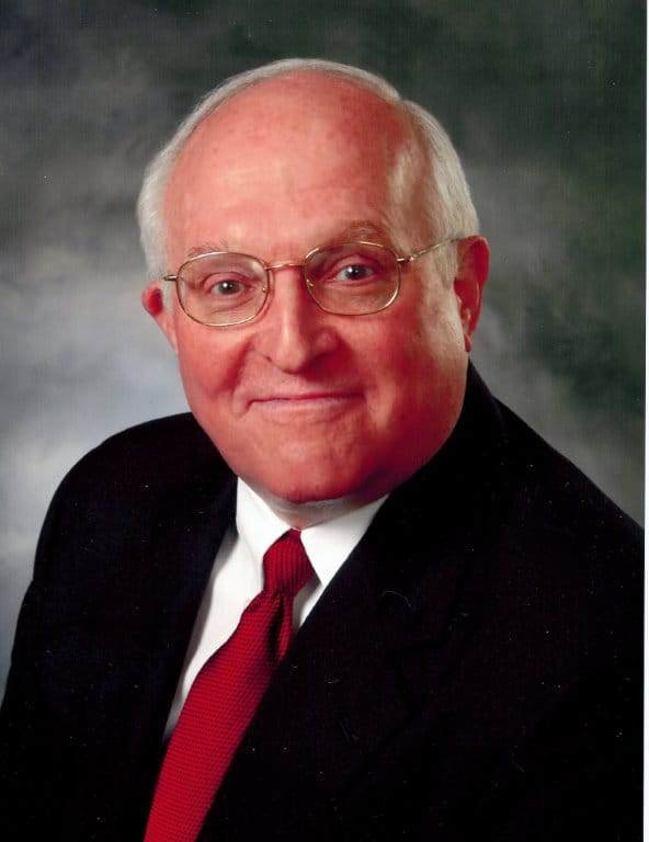 An older man with glasses, a suit, white shirt, and red tie poses in front of a neutral studio background.