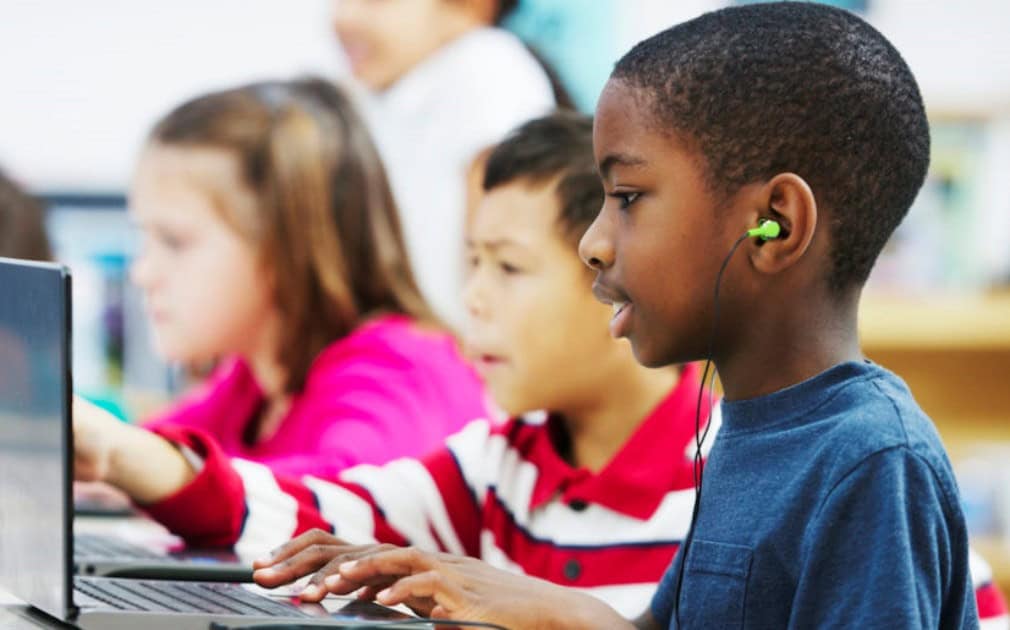 Several children sit side by side in a classroom, working on laptops—a lively scene of children's education in action.