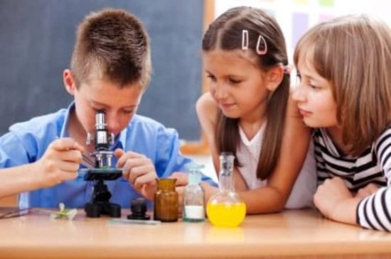 Three students observe a microscope and laboratory glassware, exploring science according to the New Science Standards in a classroom setting.