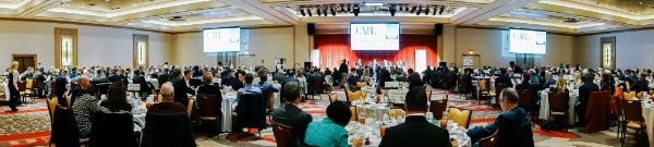 A large conference room filled with people at round tables, attending the CABL Annual Meeting and watching speakers on stage with big screens.