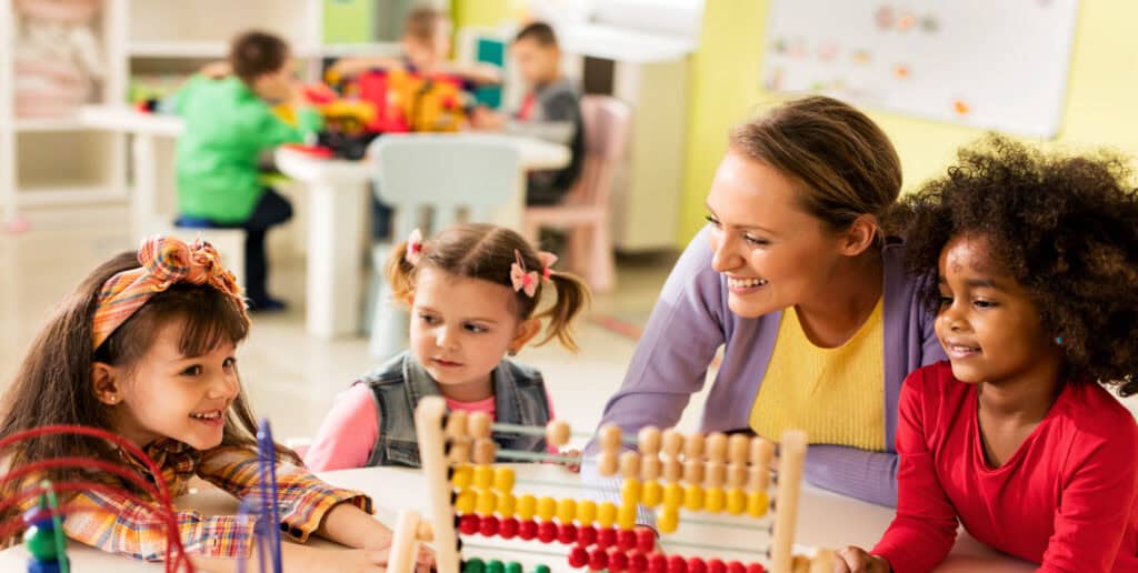 A teacher engages three young girls with an abacus, fostering early education in a brightly lit classroom filled with children.