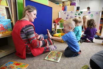 A woman sits on the floor giving a high-five to a young boy, celebrating early education in a classroom with other children and toys nearby.