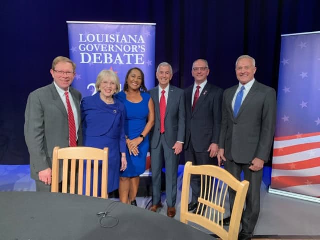 Six people in business attire smile before "Louisiana Governor's Debate" banners, ready to share their visions with voters in a studio setting.