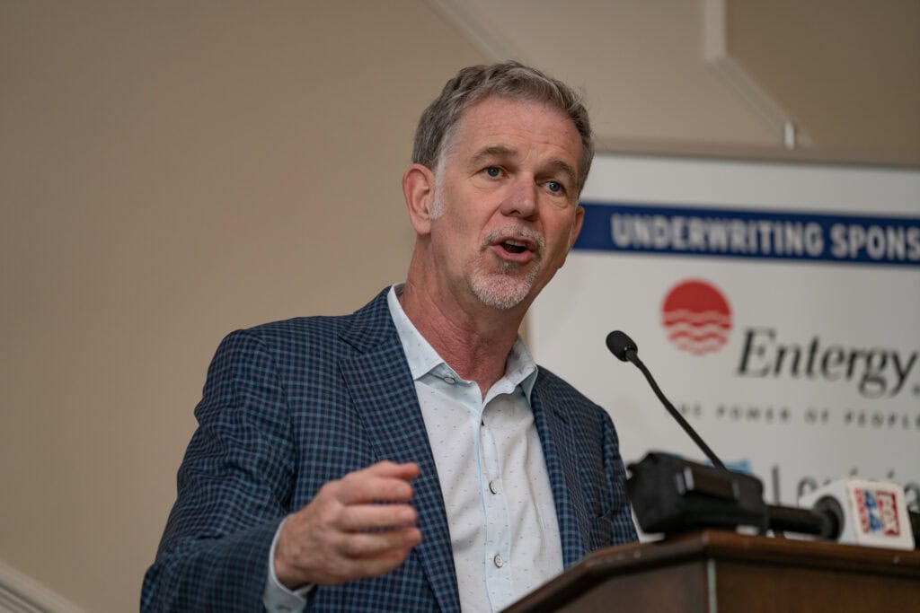 A man in a blue checked blazer, resembling Netflix CEO Reed Hastings, speaks at a podium with a sponsorship sign in the background.