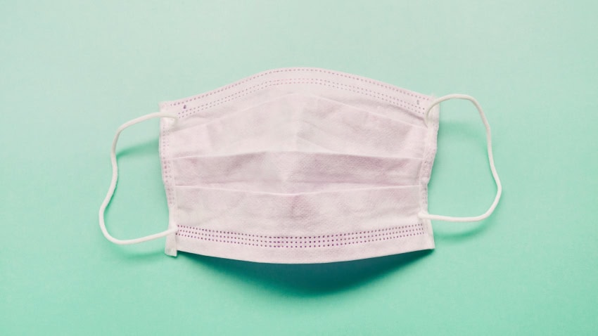 A white disposable face mask with ear loops reminds us to Stop the Spread on a light mint green background.