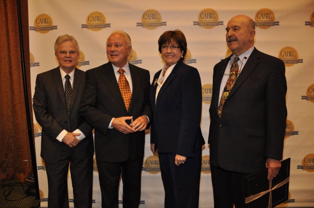 Four people in business attire stand before a "CARH" banner, honoring Governor Mike Foster's education legacy at the event.