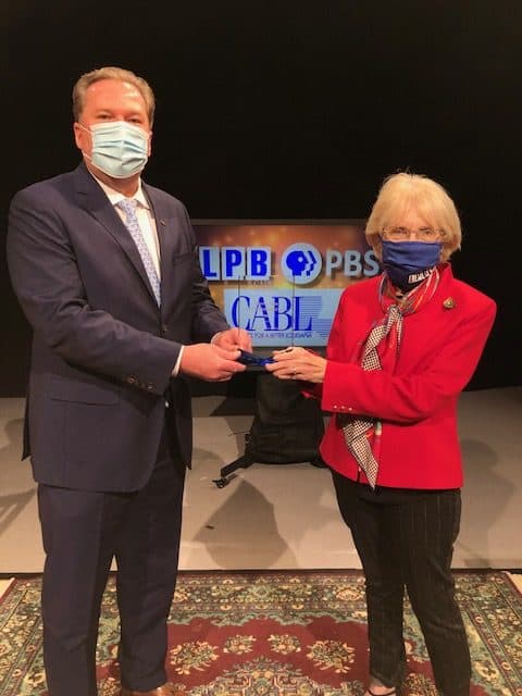 Two people wearing masks stand on a rug, holding a Distinguished Service award in front of an LPB PBS, CABL, and Beth Courtney sign.