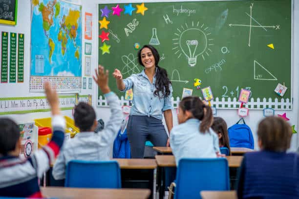 A teacher stands in front of a chalkboard, smiling and pointing as students with raised hands sit at desks, addressing teacher shortages.