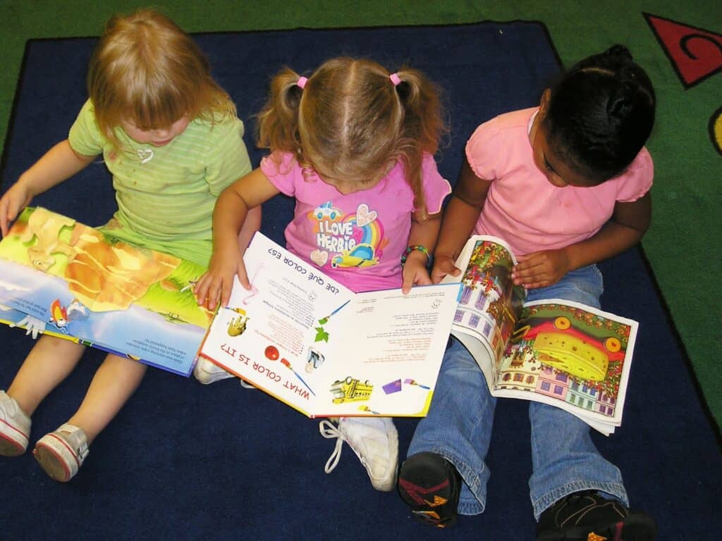 Three young children sit on a blue carpet, each reading a colorful picture book, improving reading and early reading skills together.