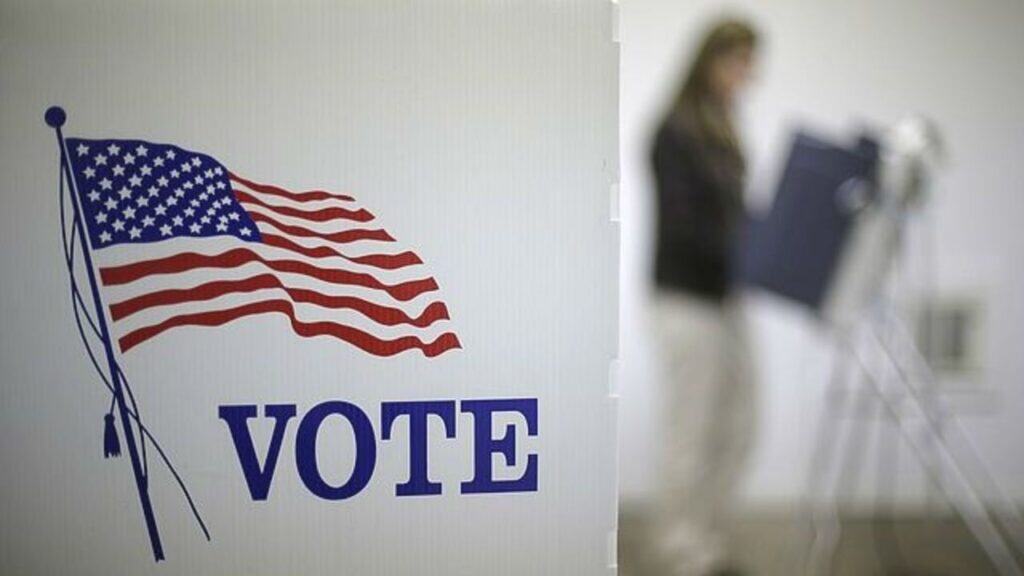 A voting booth with "VOTE" on it and an American flag, highlighting Election Focus as a person stands at a state election station in the background.