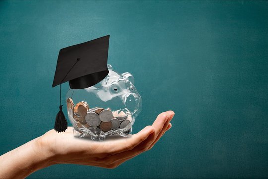A hand holds a glass piggy bank filled with coins and a graduation cap, symbolizing TOPS Scholarships, against a solid teal background.