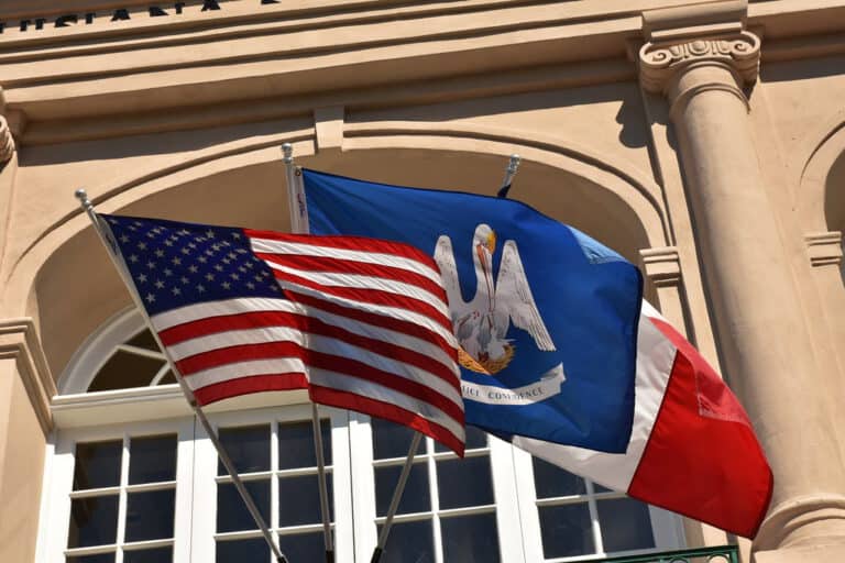 United States and Louisiana state flags fly in front of a beige building with arched windows and columns.