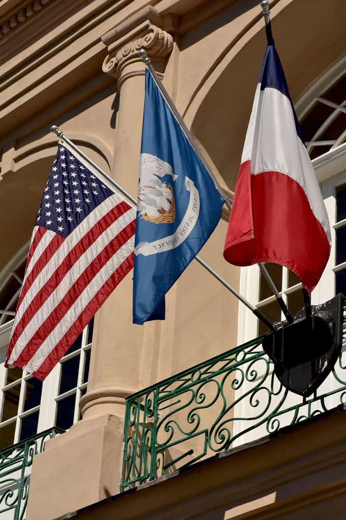 Three flags—United States, Louisiana state, and France—are mounted on the side of a beige building with a decorative green balcony.