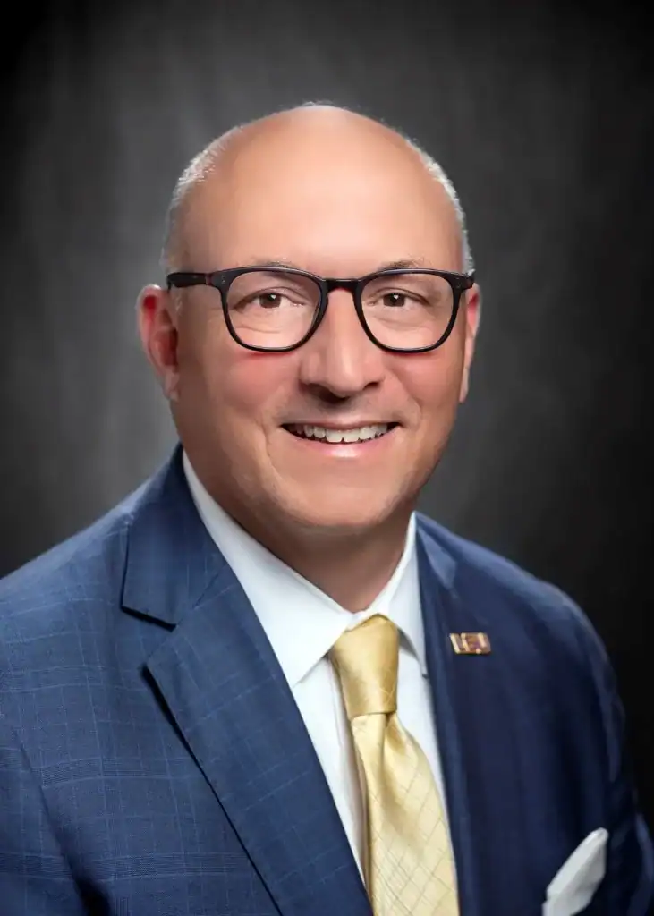 Dr. Matt Lee, bald and wearing glasses, smiles in a blue suit and yellow tie before a dark background at LSU Agricultural Center.