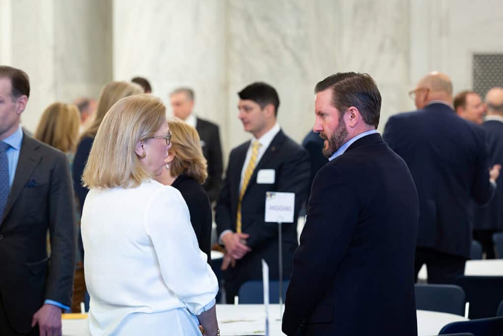 Two people are talking at a formal event, with other attendees standing and conversing in the background.