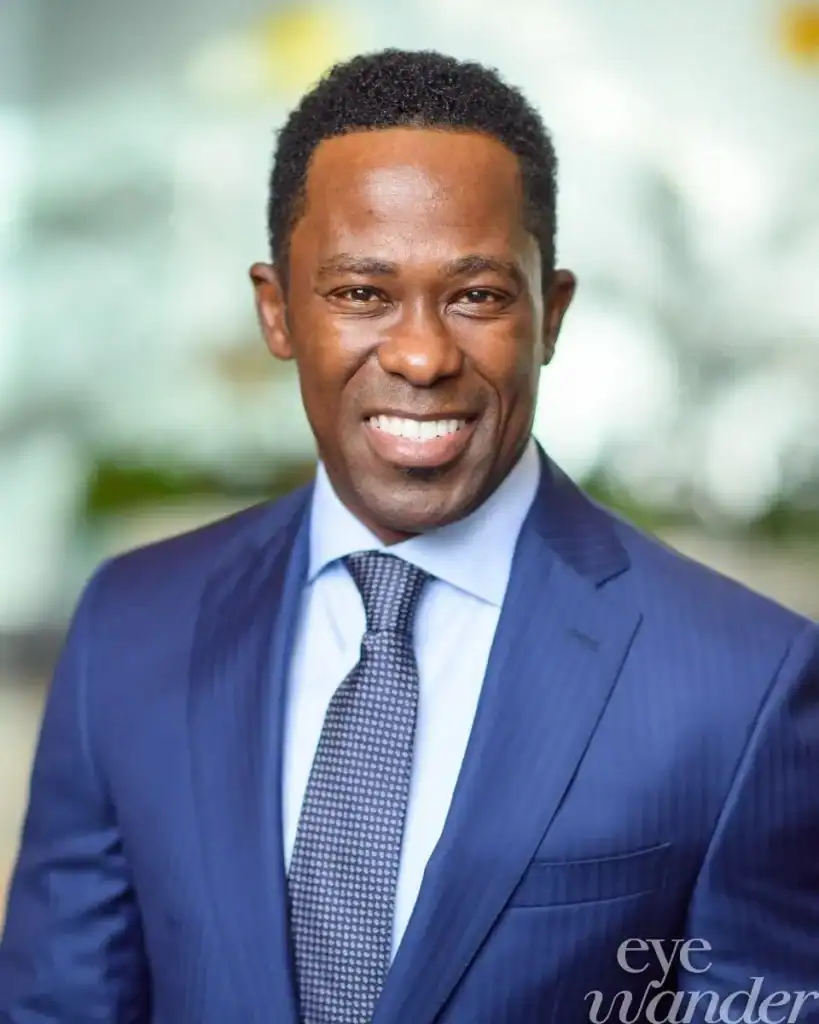 A man in a blue suit and tie smiles at the camera; "Committee of 100" appears in the corner against a blurred background.