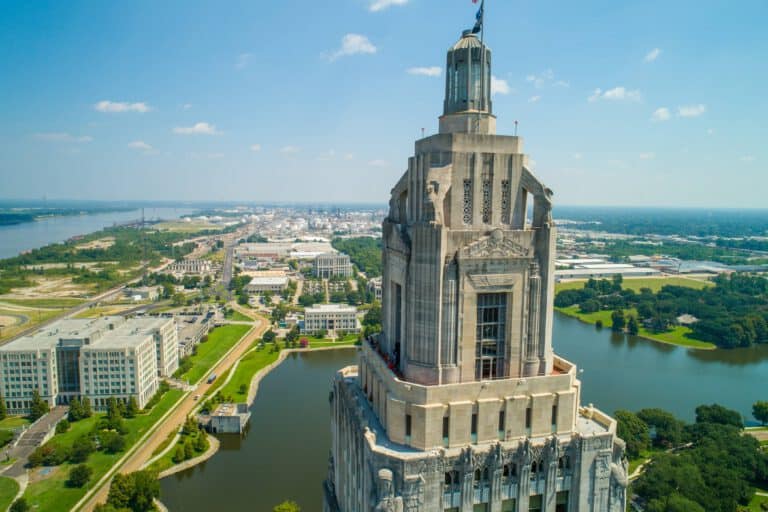 A tall, stone government building stands by a river, surrounded by greenery and other city buildings under a clear sky.