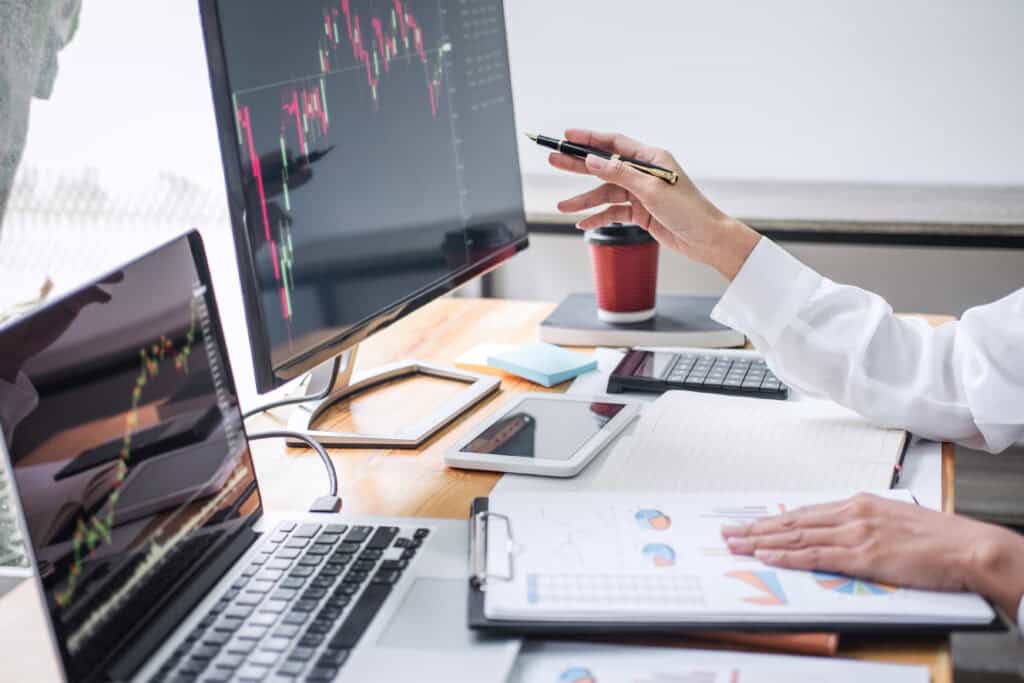 Person analyzing financial charts on monitors and papers at a desk, showcasing Leadership Louisiana skills with coffee, laptop, tablet, and calculator.