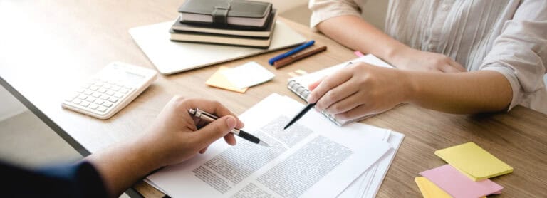 Two people sit at a desk reviewing documents, showcasing Leadership Louisiana skills with notebooks, a calculator, and sticky notes on hand.