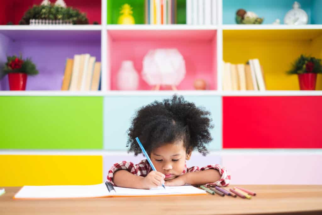 A young child writes in a notebook at a desk with colored pencils, inspired by Leadership Louisiana, surrounded by colorful shelves and books.