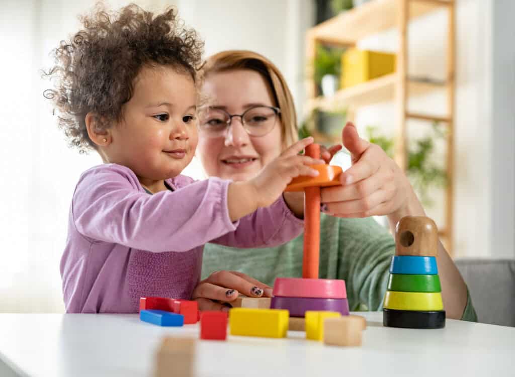 A young child and an adult stack colorful wooden rings at a table, demonstrating teamwork and Leadership Louisiana values amid scattered toys.