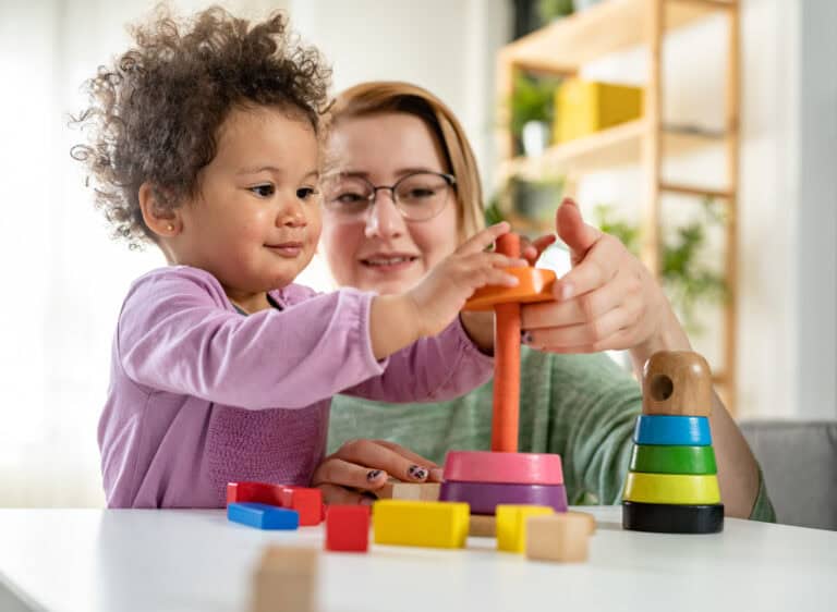 A young child and an adult stack colorful wooden rings at a table, demonstrating teamwork and Leadership Louisiana values amid scattered toys.