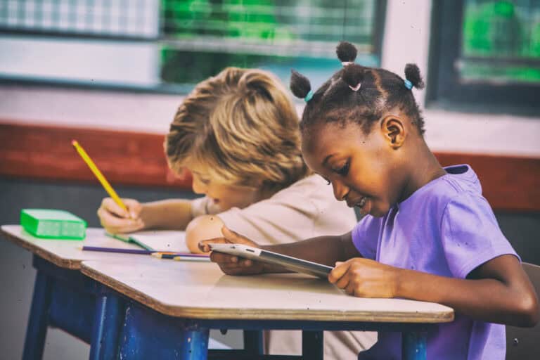 Two children sit at a classroom desk; one shows Leadership Louisiana by writing, while the other reads from a tablet device.