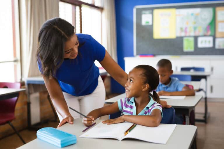 A Leadership Louisiana teacher stands by a student's desk, smiling and talking as the student writes in a notebook in class.