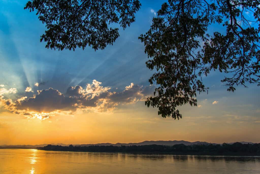 Sunset over a calm river with trees framing the top, clouds, and sun rays shining through in the background.