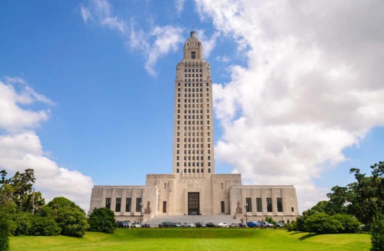 A tall, white stone capitol building symbolizes Leadership Louisiana beneath a blue sky with clouds, viewed from a green lawn.