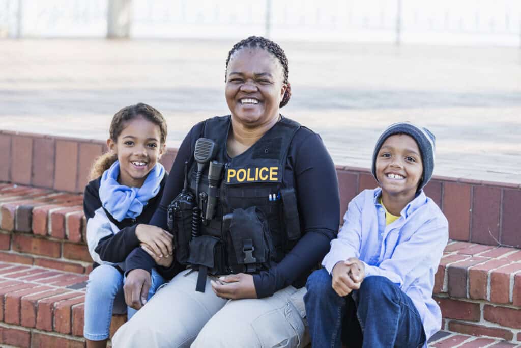 A smiling police officer sits on brick steps with two children, highlighting the human side of public safety for all.