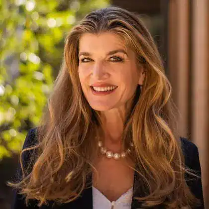Smiling woman with long wavy hair, wearing a black blazer, white top, and pearl necklace, standing outside with greenery behind her.