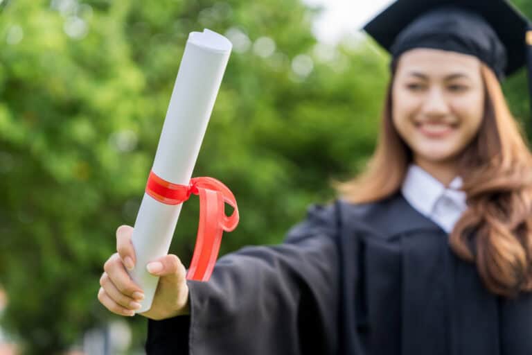 A person in a graduation cap and gown holds a diploma, celebrating Leadership Louisiana outdoors amid lush greenery.