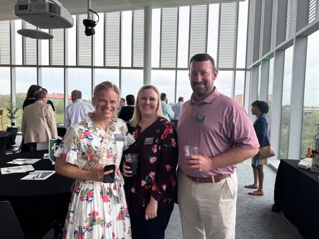 Three people stand together and smile at a social event in a bright room with large windows and other attendees in the background.