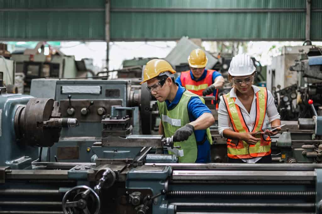 Three factory workers in safety vests and helmets demonstrate Leadership Louisiana while operating machinery inside an industrial workshop.