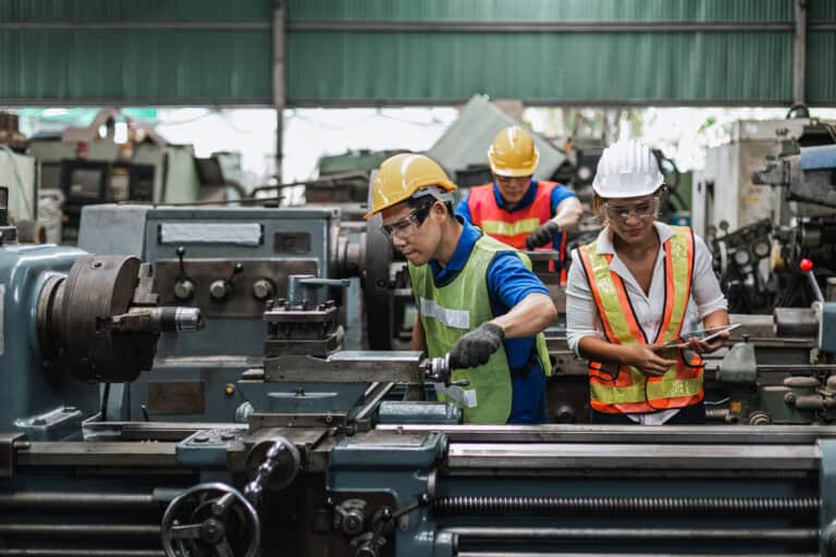 Three factory workers in safety vests and helmets demonstrate Leadership Louisiana while operating machinery inside an industrial workshop.