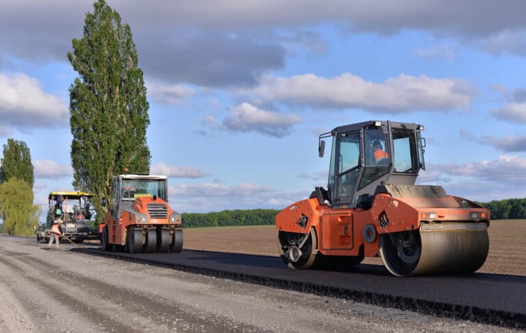 Two orange road rollers and a paver, part of Leadership Louisiana's efforts, lay new asphalt on a rural road under a partly cloudy sky.