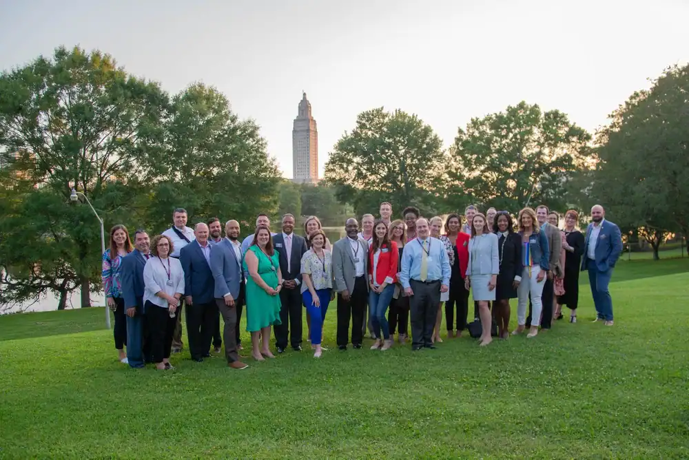 A group of people poses on a grassy lawn with trees and a tall building in the background at sunset.