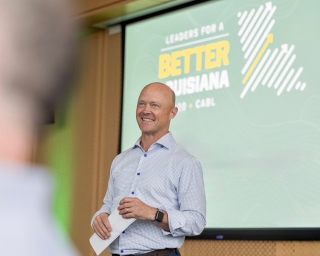 A man stands and smiles while holding a paper, in front of a screen displaying 