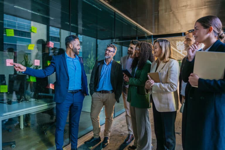 A group of six professionals stands by a glass wall with sticky notes, as a Leadership Louisiana member gestures and speaks to the others.