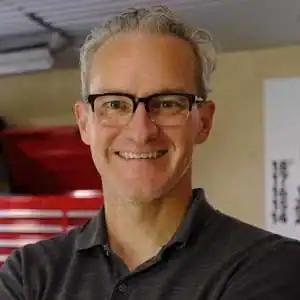 A man with short gray hair and glasses stands indoors, smiling, wearing a dark collared shirt.