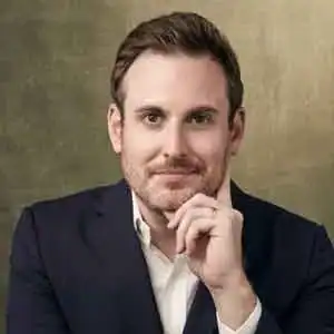 Man in a dark suit and white shirt rests his chin on his hand, looking at the camera, with a neutral background.